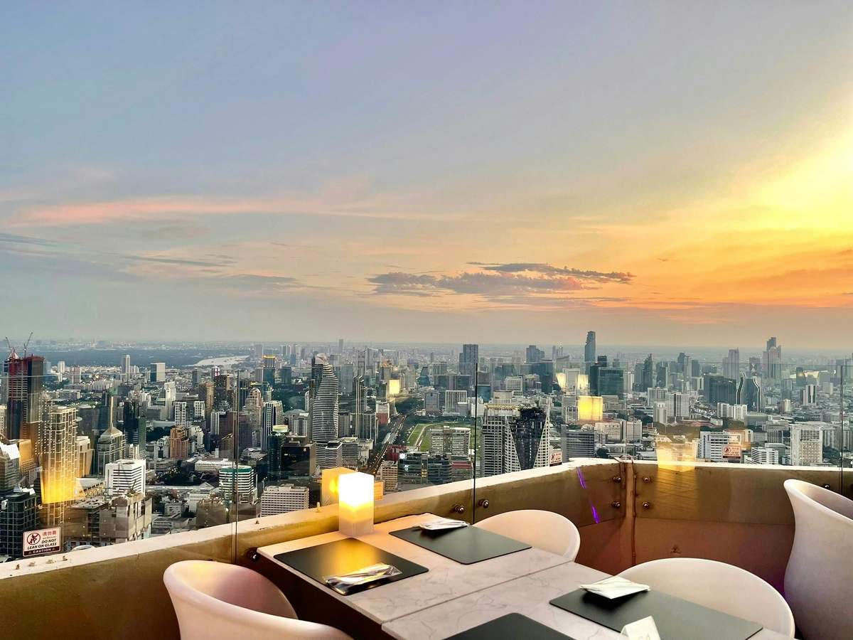 Restaurante na cobertura ao pôr-do-sol, com vista para o horizonte da cidade, com mesas postas e um brilho suave proveniente das lanternas nas mesas.