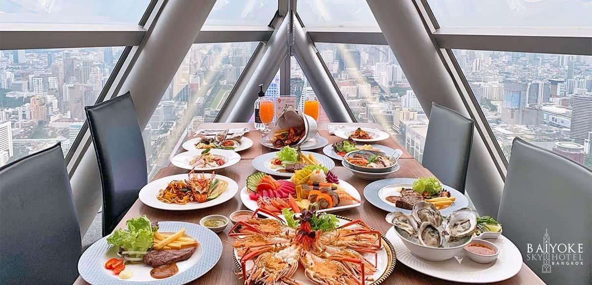 Bangkok: Buffet Lunch/Dinner at Baiyoke Sky Restaurant A luxurious spread of seafood and gourmet dishes on a table with a cityscape view through large triangular windows.