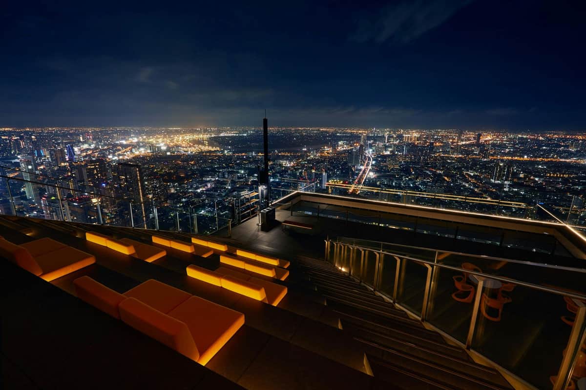 スカイビーチマハナコンバンコク:専用スカイビーチルーフトップナイト(午後7時〜午後11時) 夜には街並みを一望できる屋上シーティングエリアで、遠くには照明付きの建物や通りがあります。