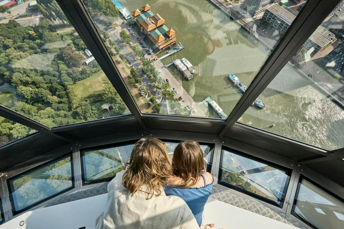 Euromast + Remastered Two individuals, viewed from behind, observing a waterfront and boats from a high vantage point through large windows.
