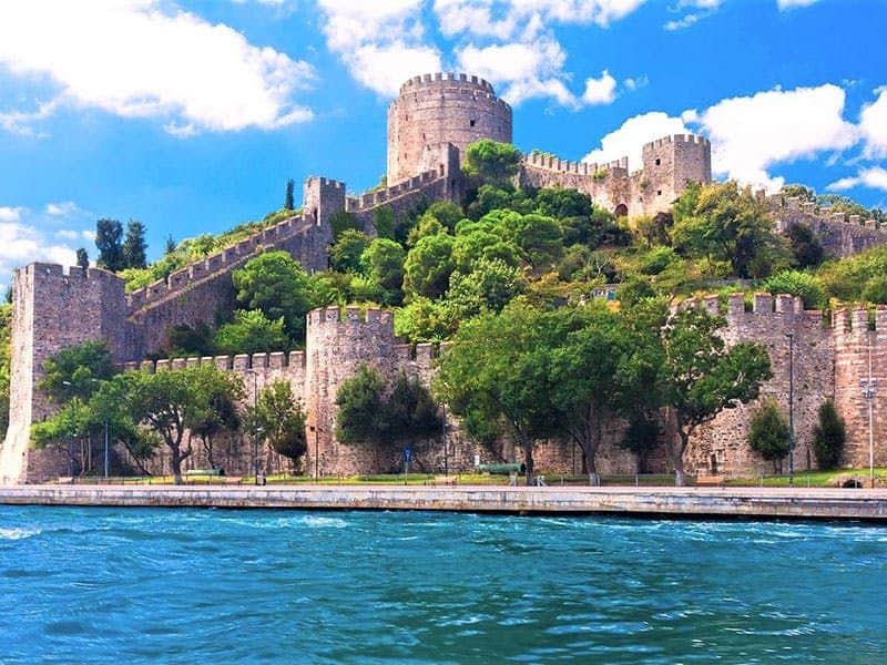 Bosphorus, Black Sea & Dolmabahce Palace: Boat Tour with Lunch + Entry A historic stone fortress with towers and walls on a hill, surrounded by greenery, adjacent to a body of water.