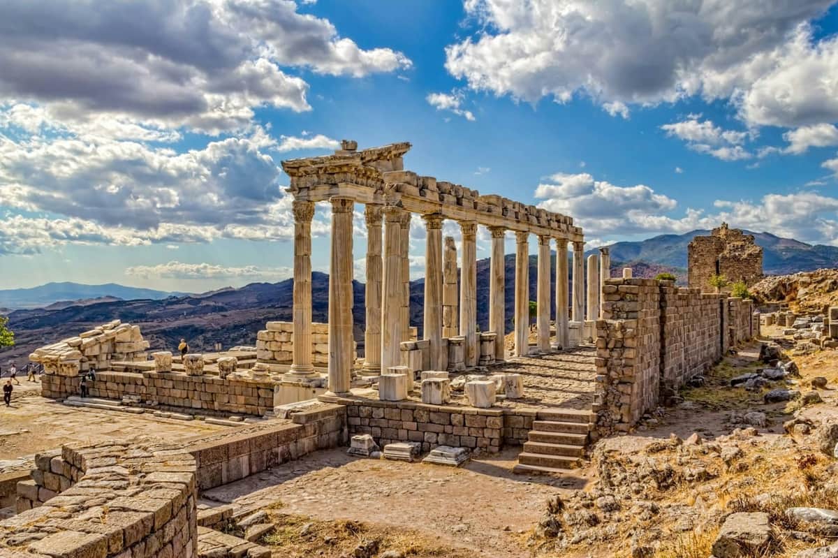 Ancient ruins with stone columns and arches under a partly cloudy sky with mountains in the background.