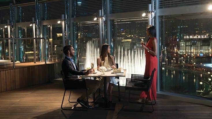 Burj Khalifa: Dining Experience Three people dining on a balcony at night with a cityscape and illuminated fountain in the background.