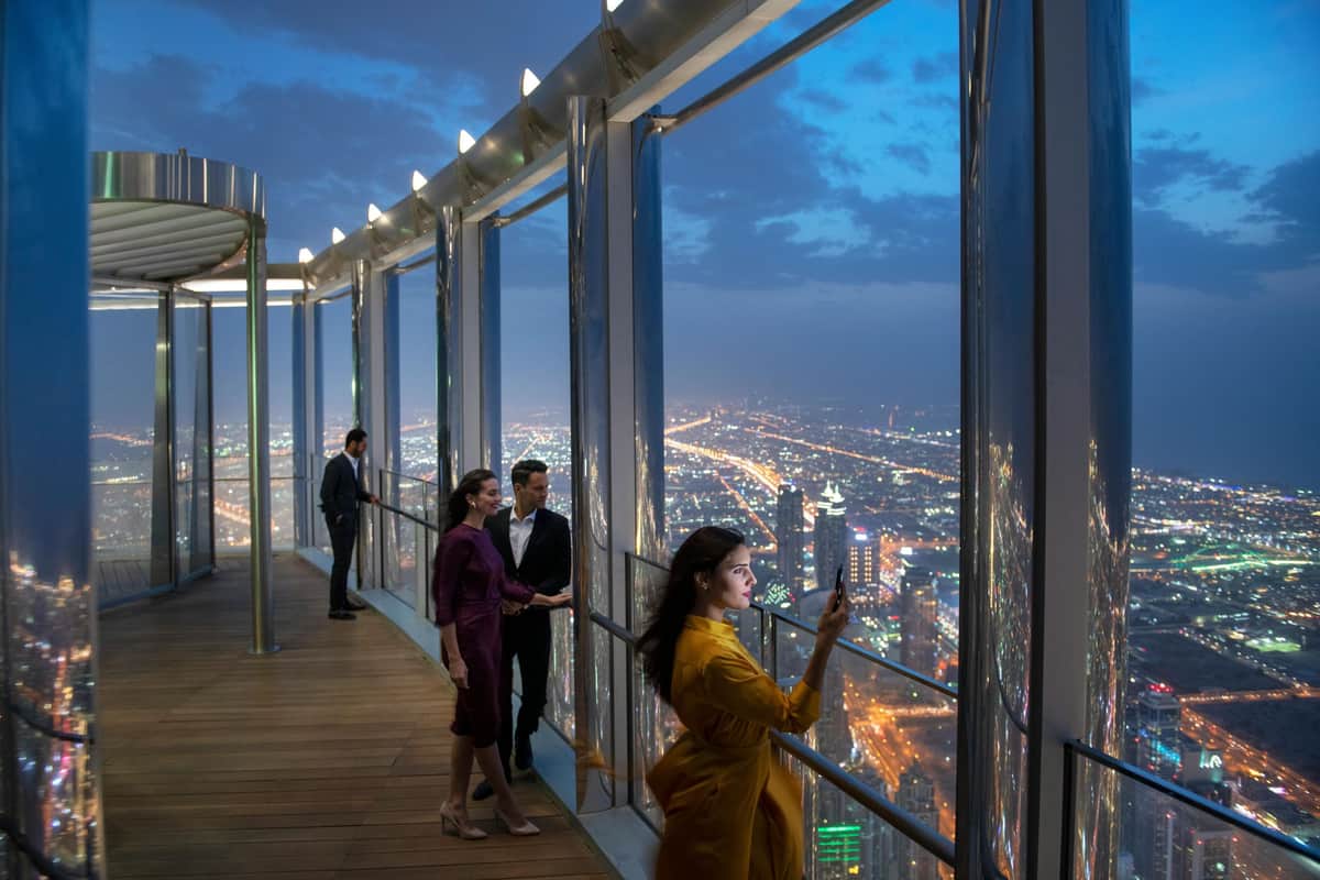 Burj Khalifa: The Lounge Level 154 Ticket People standing on an observation deck at night, overlooking a brightly lit cityscape through large glass windows.