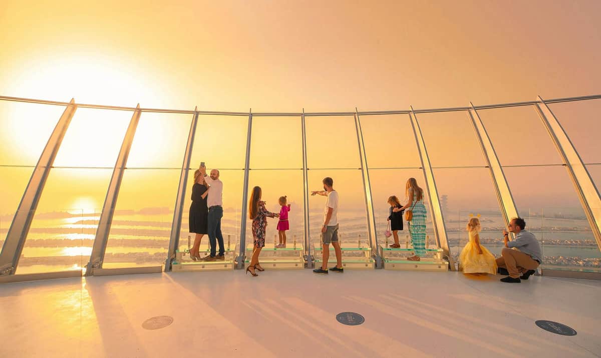 The View at The Palm: Entry Ticket (Level 52) People standing and admiring the view through large windows on an observation deck at sunset, with orange sunlight filling the scene.