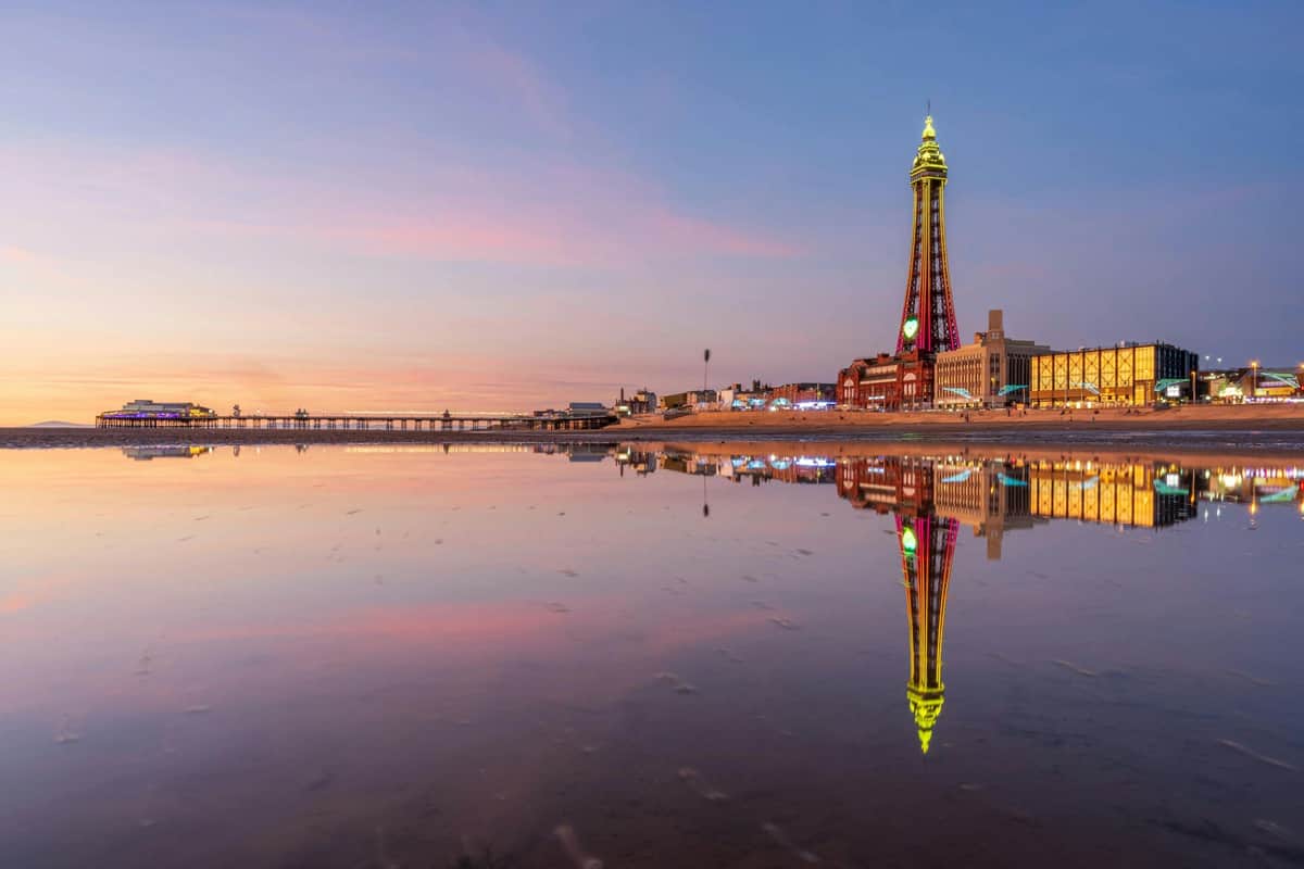 The Blackpool Tower: Tower Top Entry Ticket + 4D Cinema A tall, illuminated tower and surrounding buildings reflected in a calm body of water at sunset.