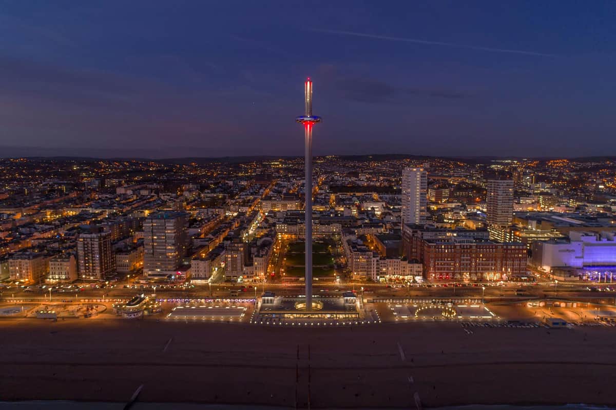 Brighton i360: City Lights Flight + Welcome Drink A cityscape at dusk featuring a tall observation tower with lights, surrounded by buildings and illuminated streets.