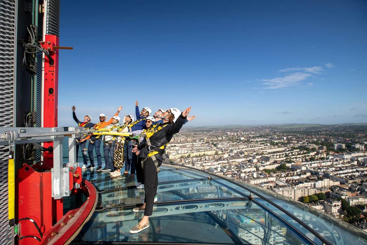 Brighton i360: Sky Walk Experience A group of people in safety harnesses lean out from a glass platform atop a tall building, overlooking a cityscape.