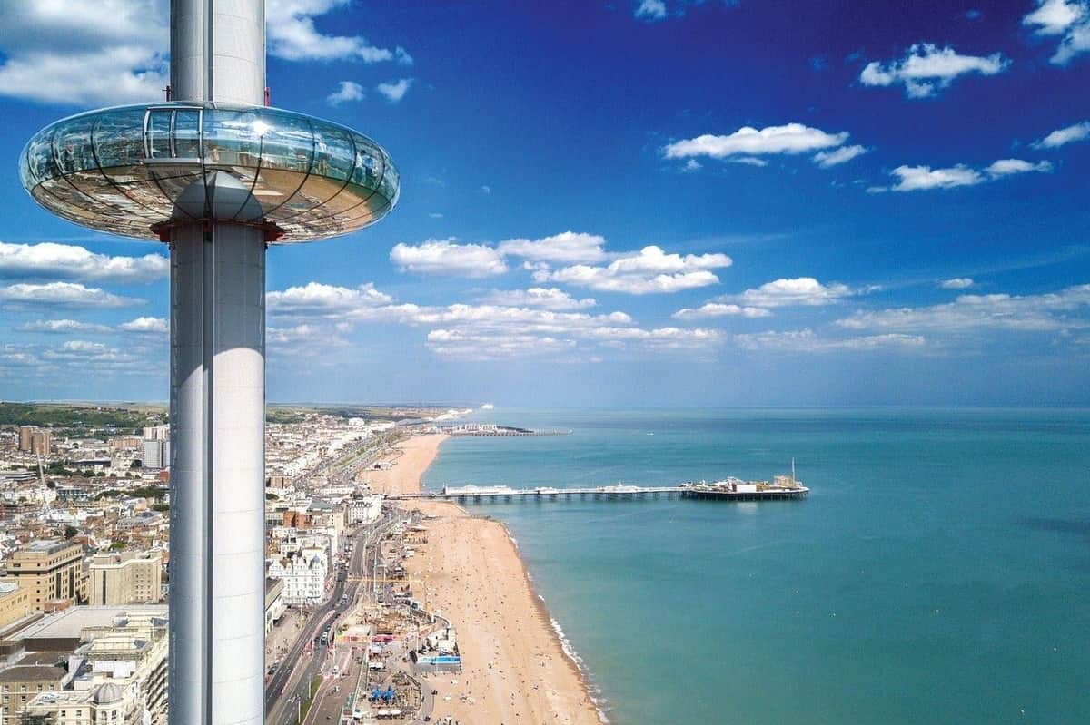 Brighton i360: View 360 Anytime Ticket A seaside cityscape with a beach, pier, and a tall observation tower under a blue sky with clouds.