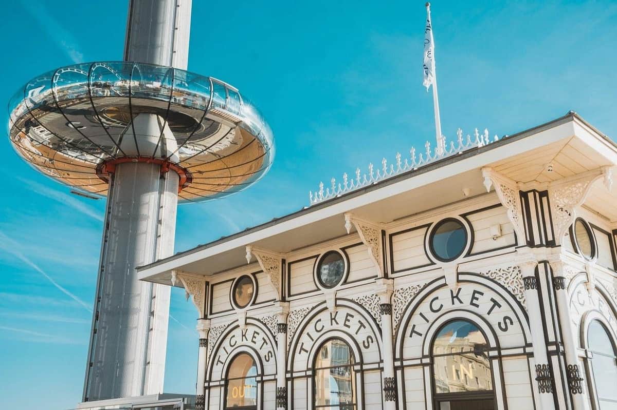 Brighton i360: View 360 Ticket A ticket booth in ornate style beside a modern tower with a circular observation pod, under a clear blue sky.