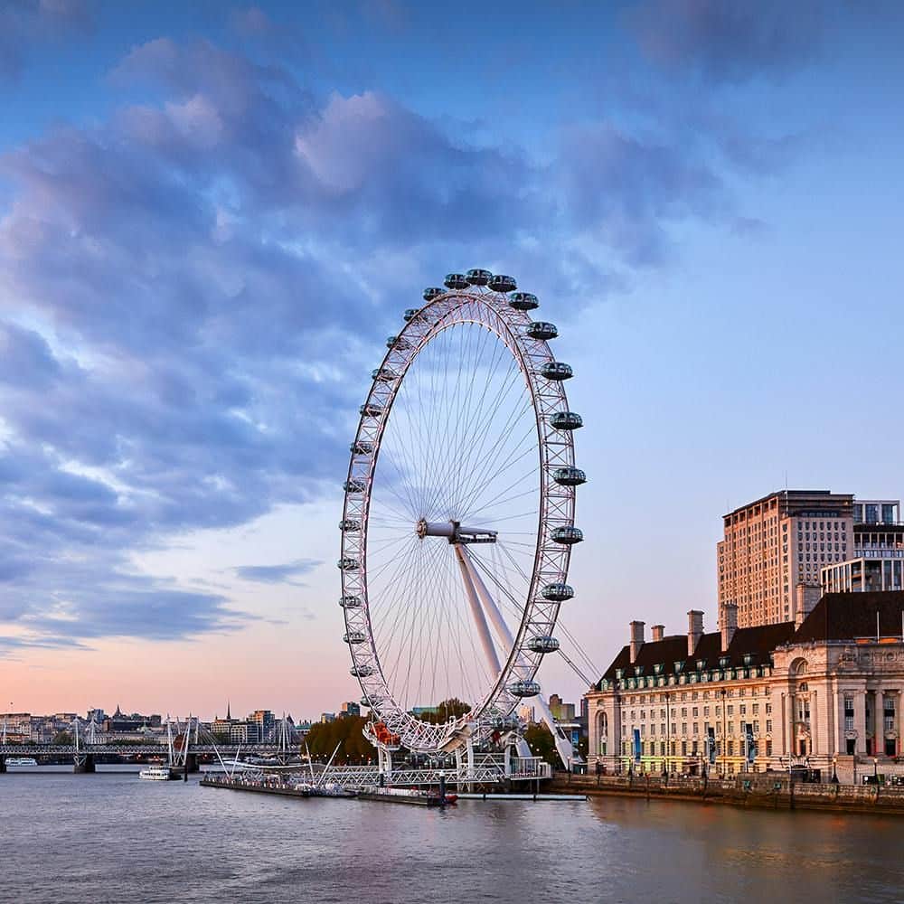 London Eye: Entry Ticket A large Ferris wheel by a riverside with nearby buildings and a bridge under a partly cloudy sky at sunset.