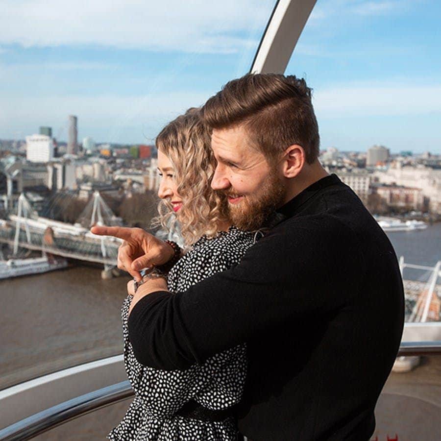 London Eye: Entrada de acceso rápido + Vaina privada Una pareja se abraza en el interior de una noria, con vistas a un río y a un paisaje urbano con puentes y edificios al fondo.