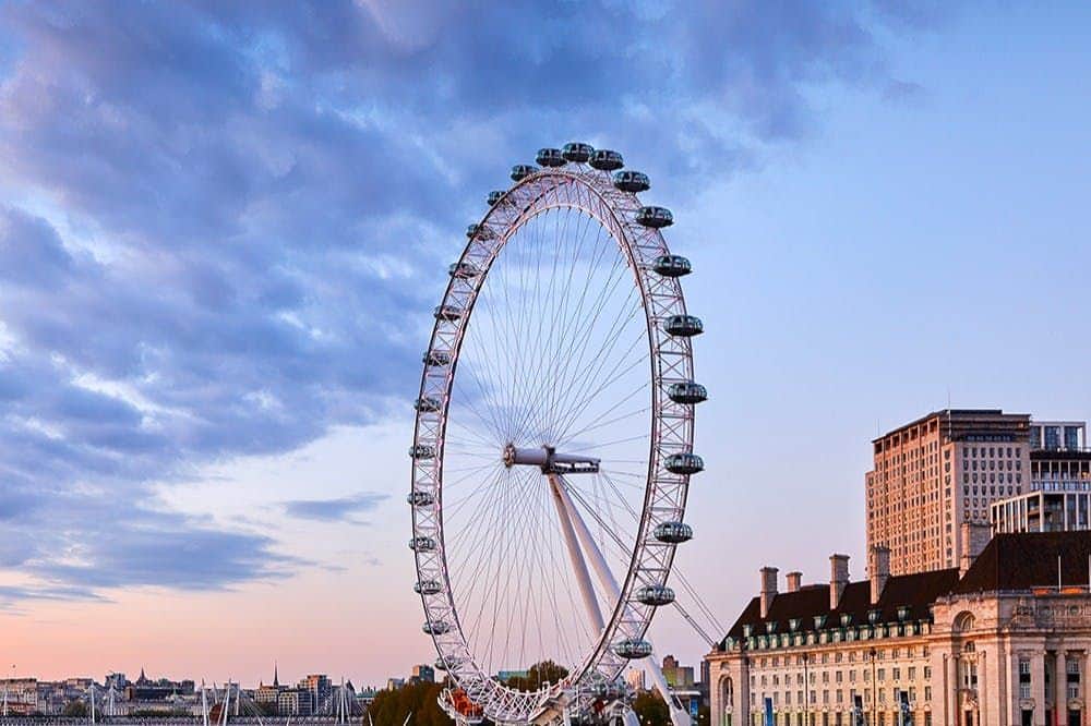 London Eye, Madame Tussauds & SEA LIFE London: Entry Ticket A large Ferris wheel, known as the London Eye, against a partly cloudy sky with buildings in the background at sunset.