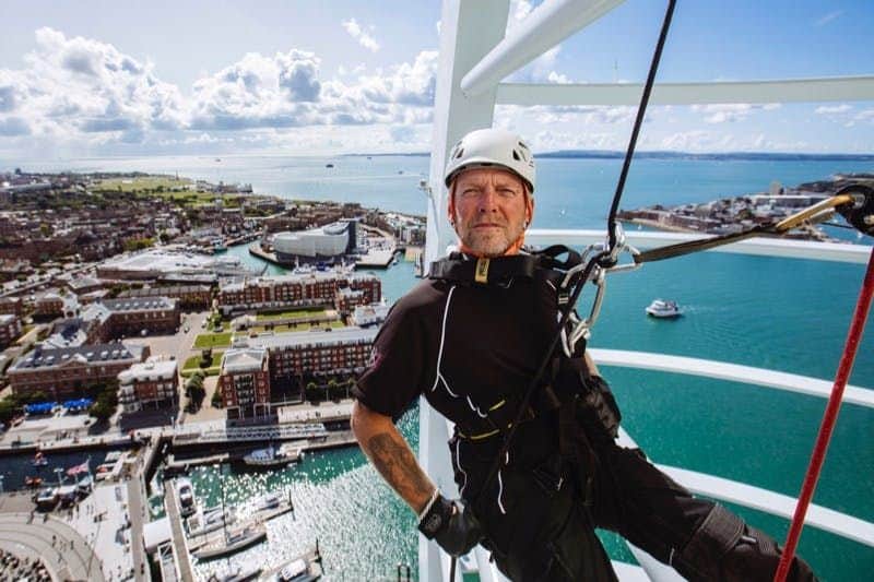 The Spinnaker Tower: Entry Ticket A man in climbing gear and helmet is suspended from a structure high above a marina and coastal cityscape.
