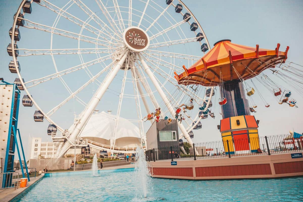 Navy Pier Centennial Wheel: Entry Ticket Ferris wheel and spinning carousel at Navy Pier with people riding, near a water fountain, under a clear sky.
