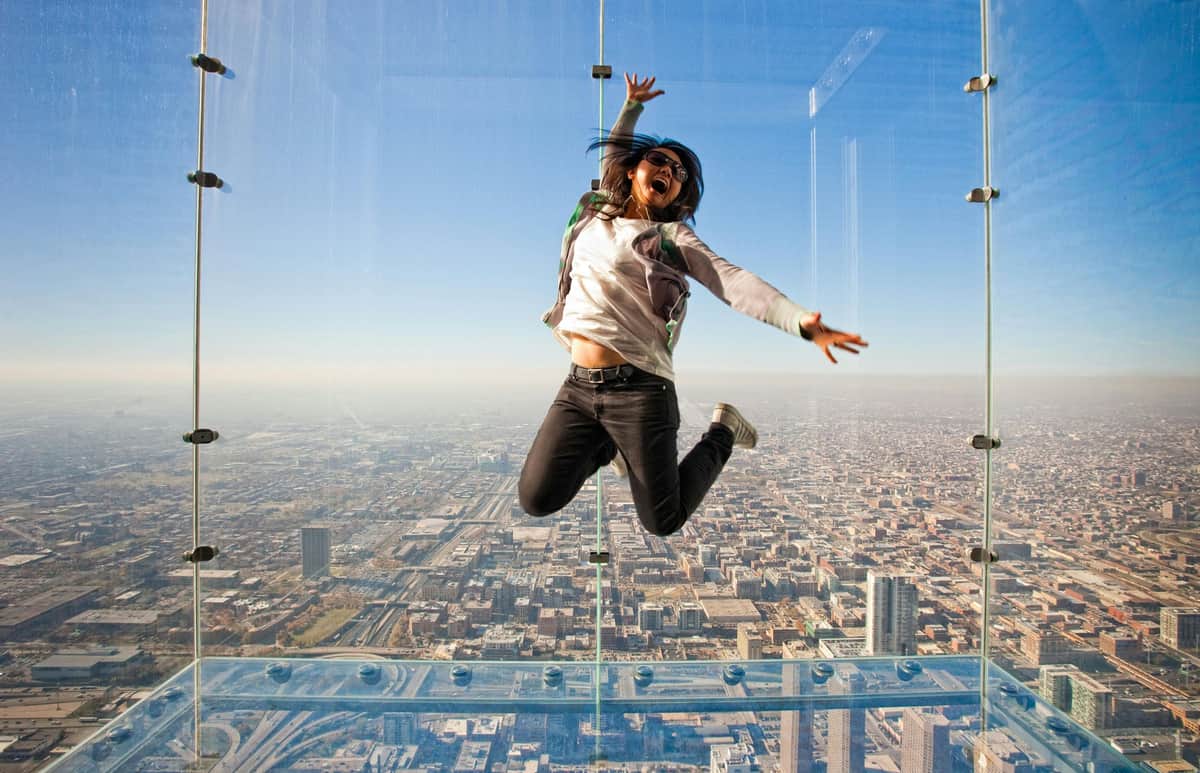 Skydeck Chicago: Entry Ticket Person jumping inside a glass observation deck, smiling with arms outstretched, with a cityscape view in the background.