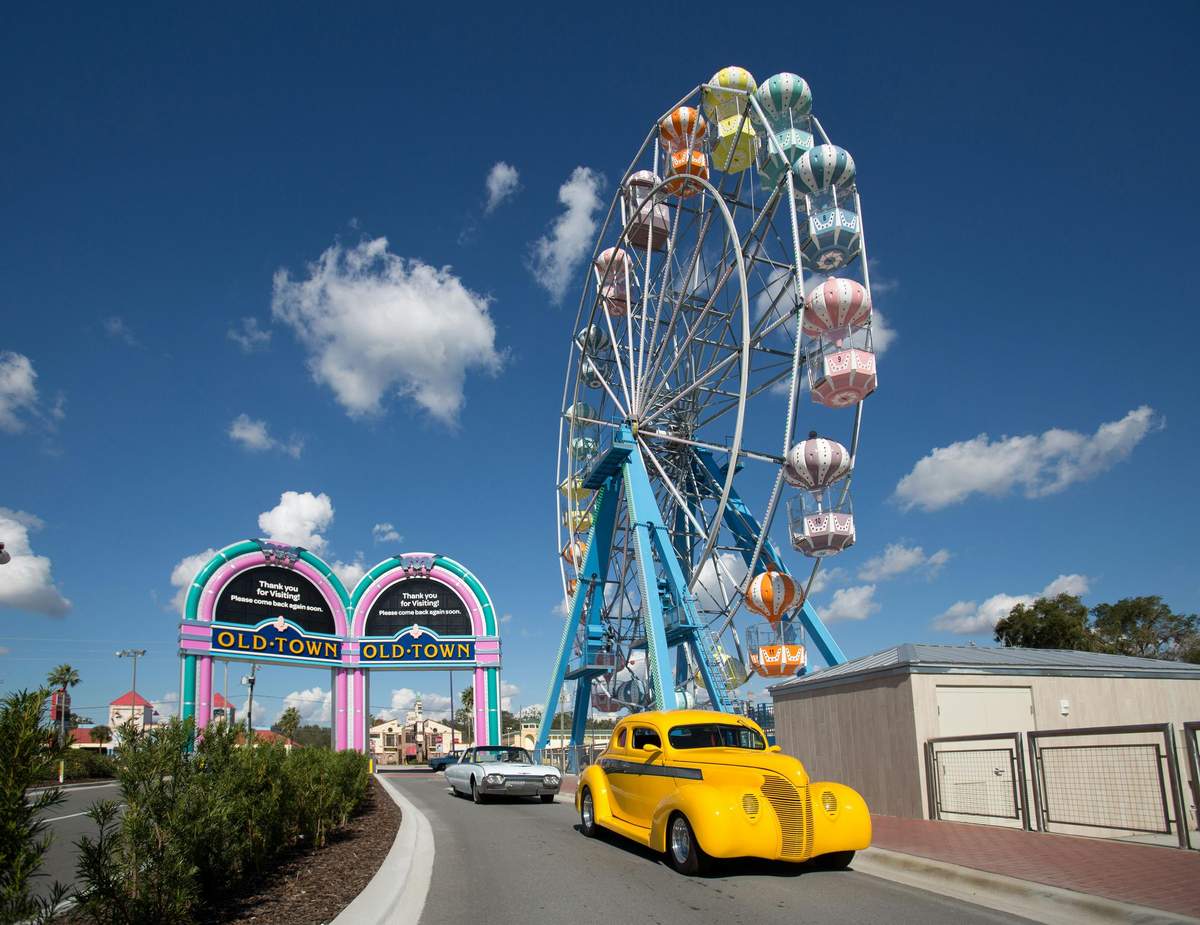 Old Town Kissimmee Ferris Wheel and Attractions Pass A colorful Ferris wheel towers over a road with a bright yellow vintage car, a grey car, and an "Old Town" entrance sign.