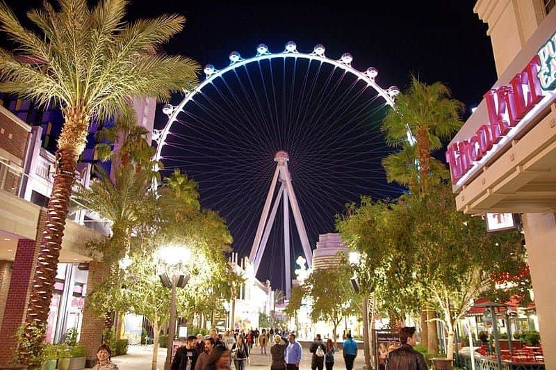 LINQ High Roller: Entry Ticket People walk along a brightly lit, palm tree-lined street at night with a large illuminated Ferris wheel in the background.
