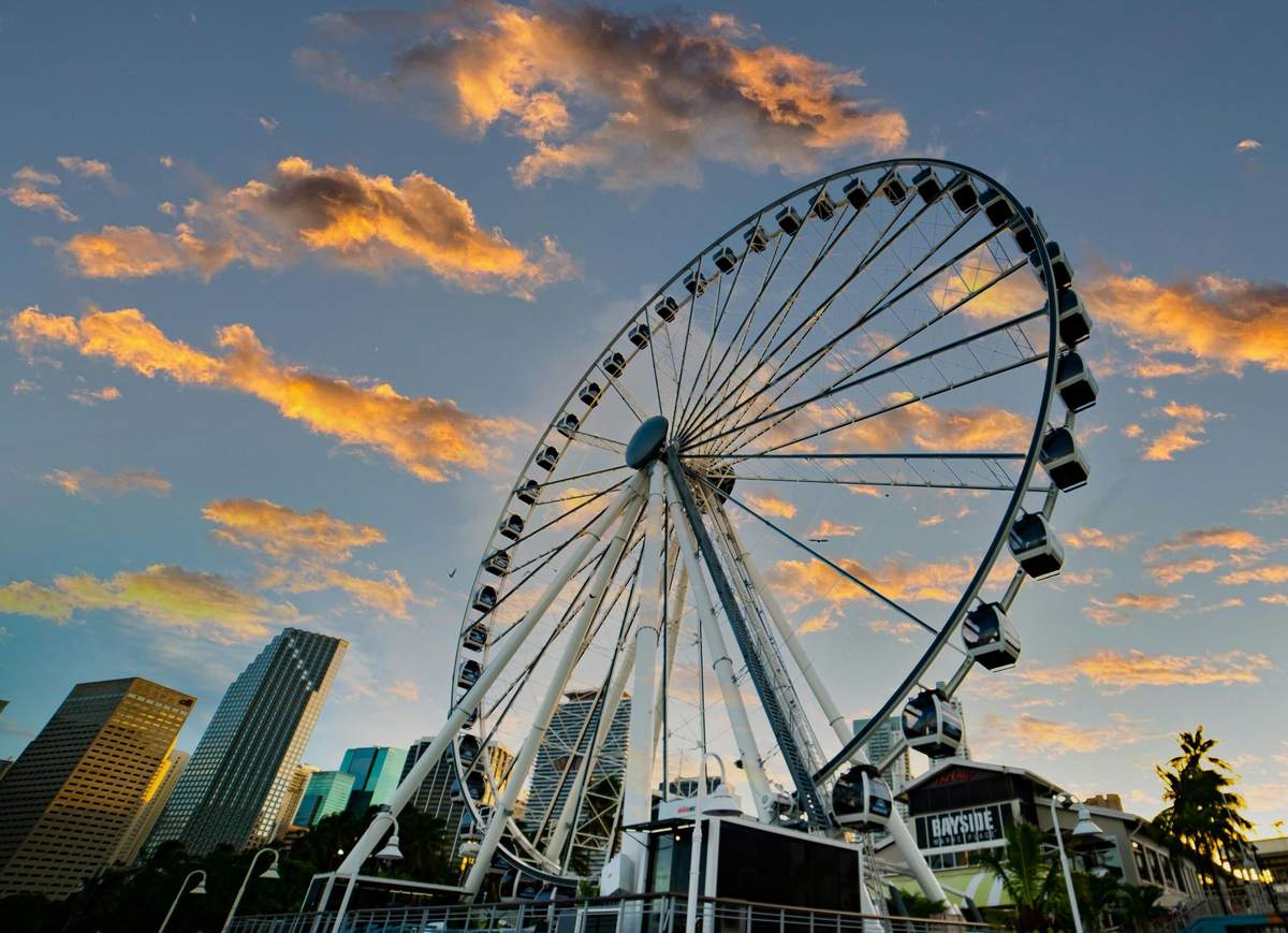Skyviews Miami Observation Wheel: Entry Ticket Ferris wheel against a sunset sky, with city buildings in the background and some clouds illuminated by the setting sun.