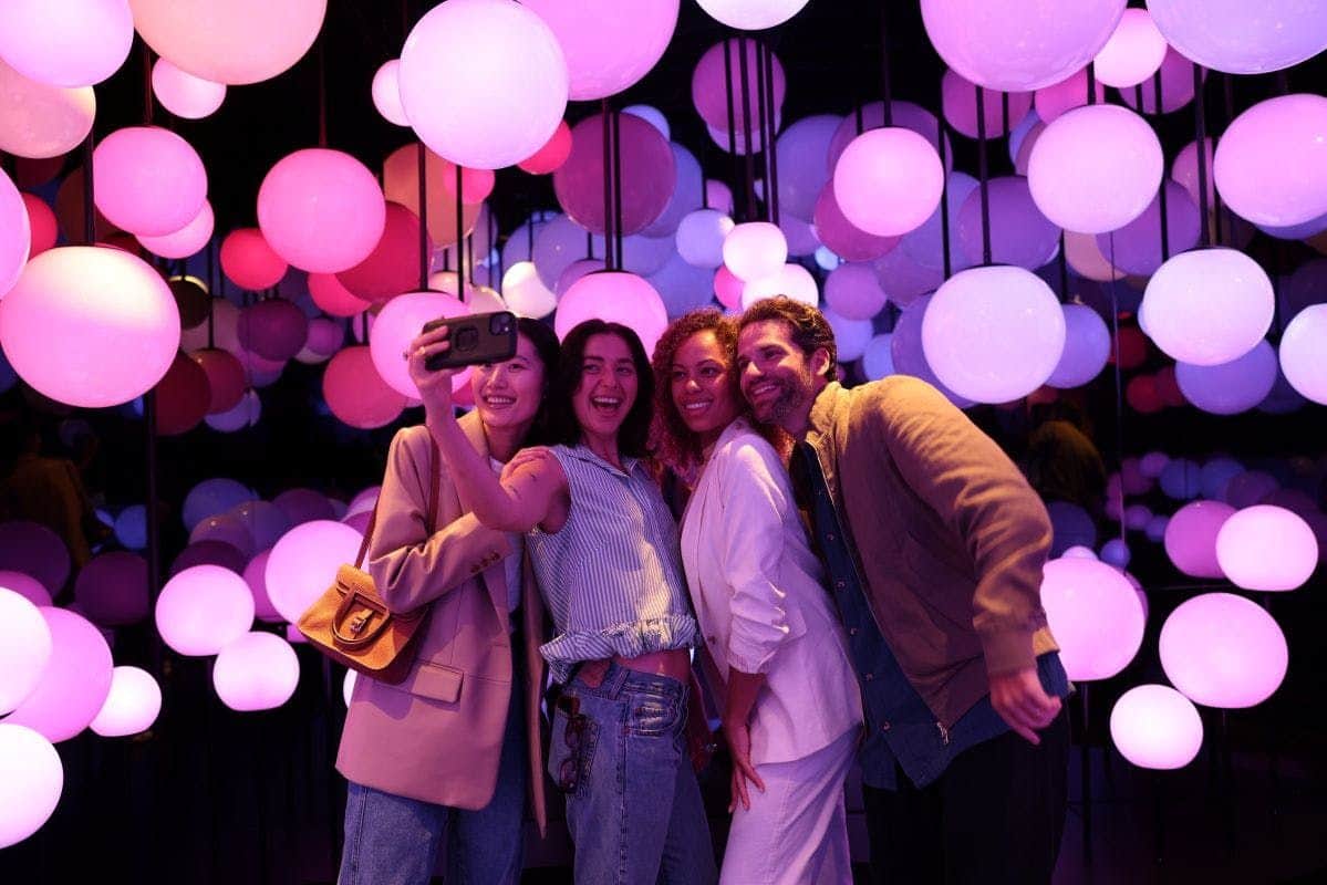 Edge NYC: Entry + Champagne Four people smiling and taking a selfie in a room filled with hanging spherical lights glowing in purple and pink hues.