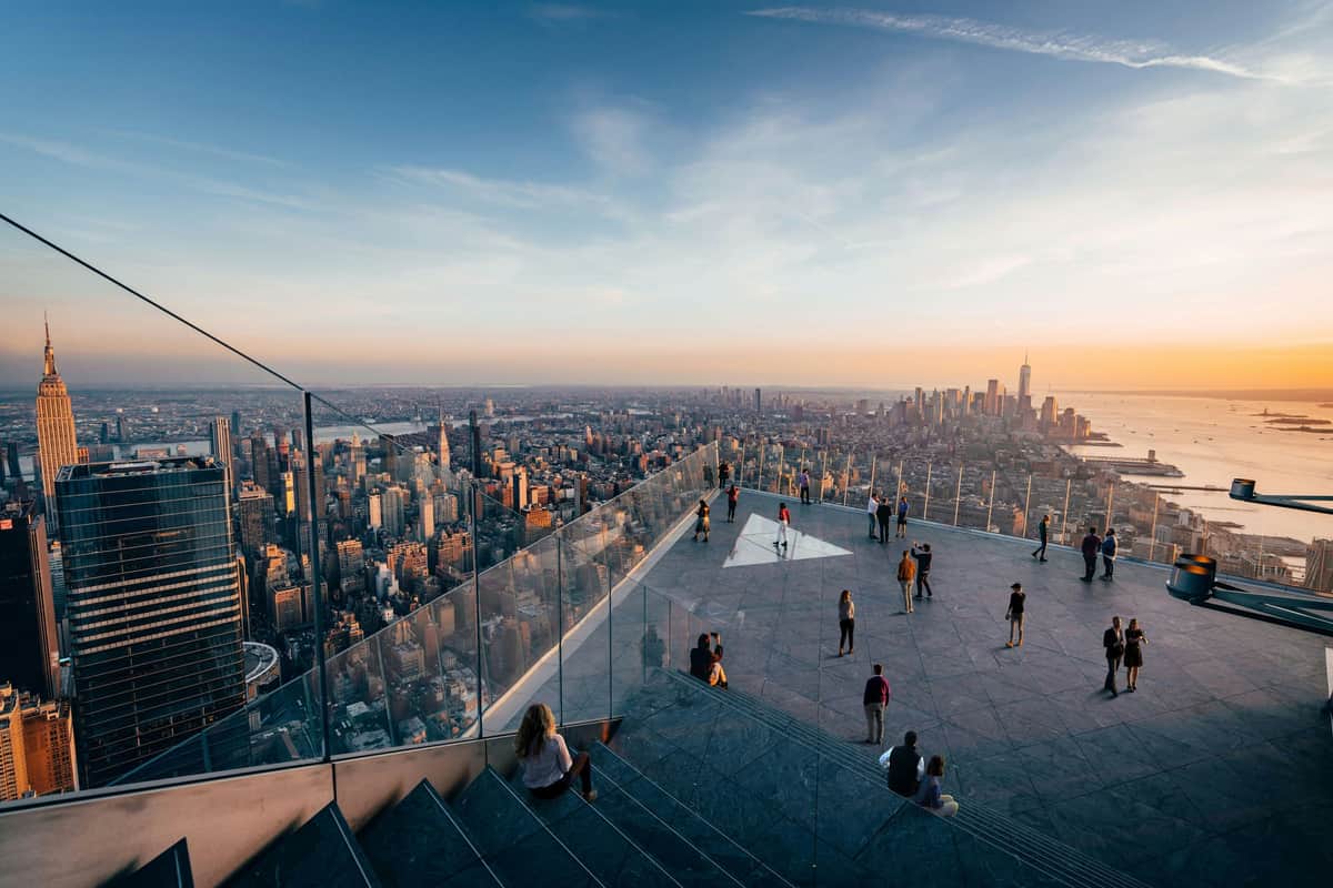 Edge NYC: Entry Ticket People standing and sitting on a large outdoor observation deck with a glass railing, overlooking the city skyline at sunset.