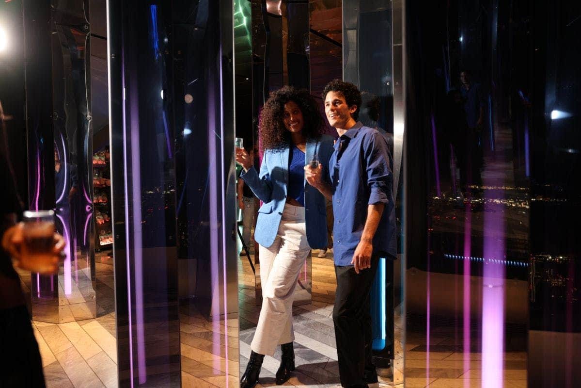 Edge NYC: Flex Pass Two people dressed stylishly holding glasses, standing in a mirrored space with city lights visible in the background.