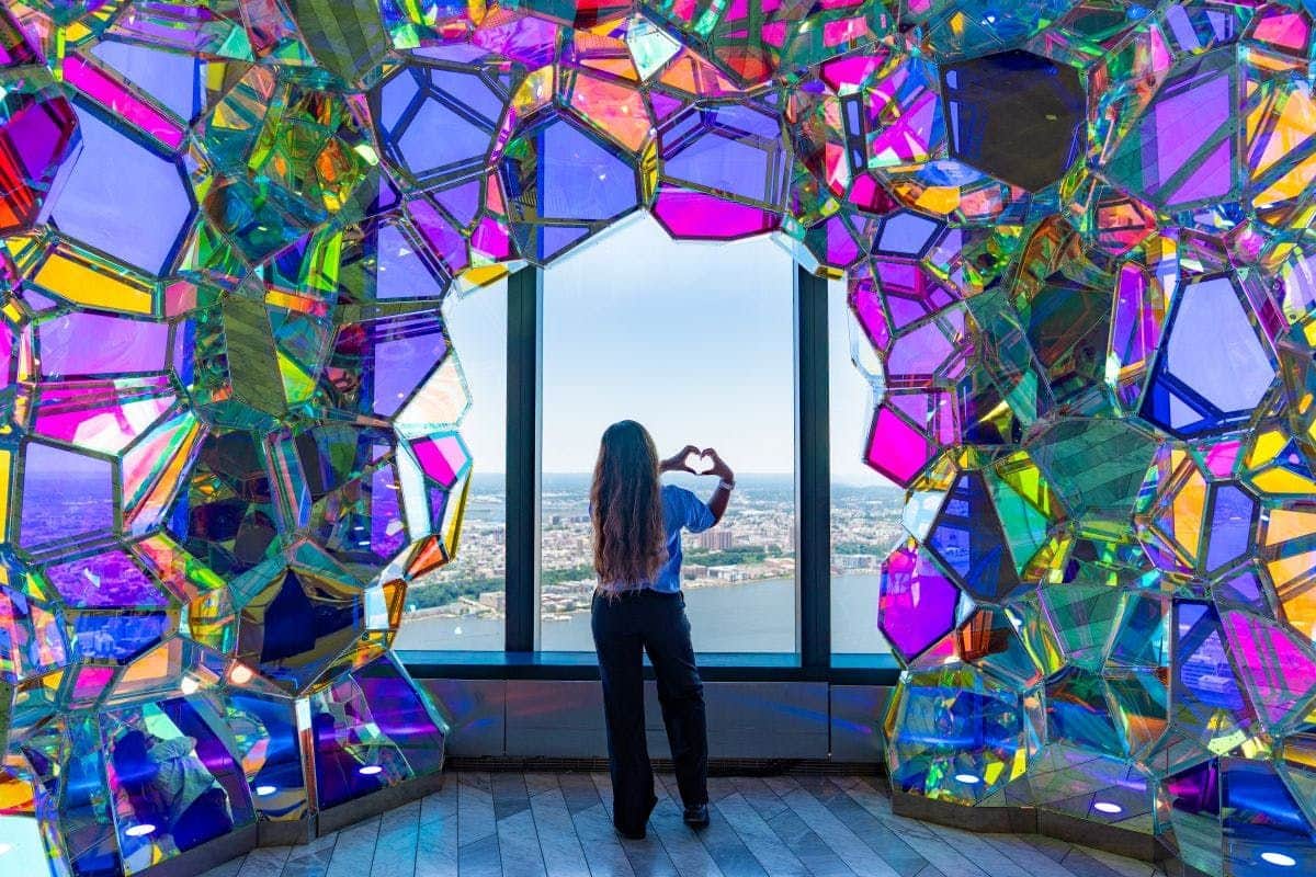 Edge NYC: Sunset VIP Admission A person stands facing a window framed by colorful, faceted panels, making a heart shape with their hands. Cityscape visible outside.