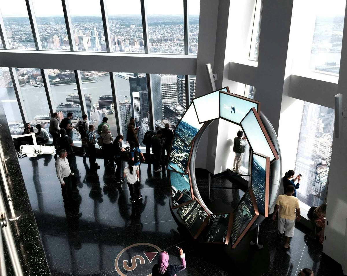 New York: 9/11 Memorial Guided Tour + One World Observatory Entry People gather at an observation deck with large windows, providing a view of a cityscape and river. A multi-screen display stands nearby.