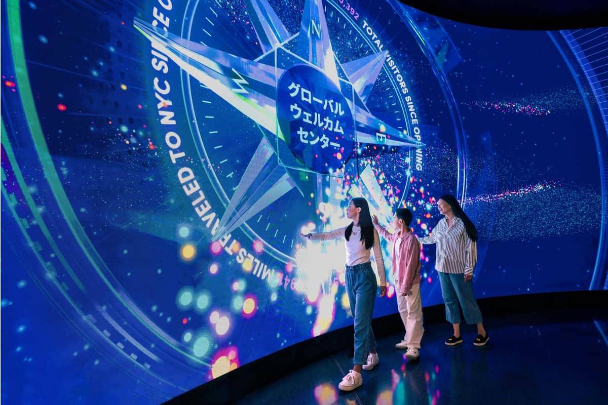 Three people interact with a large, vibrant digital display featuring a compass design and Japanese text in a dimly lit room.