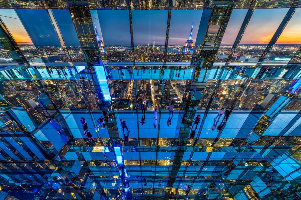 SUMMIT One Vanderbilt: Entry Ticket People observing a cityscape from within a glass-walled structure with multiple reflections and neon blue lighting.