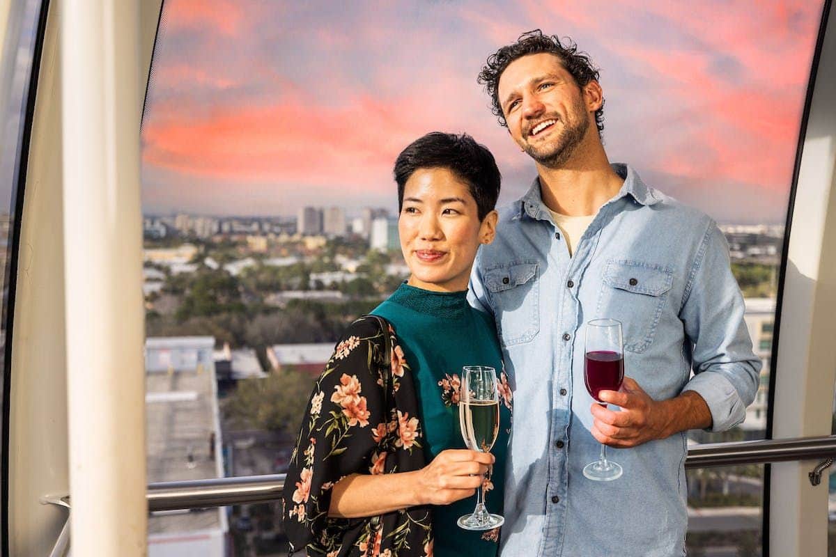ICON Park: The Orlando Eye A smiling couple stands on a balcony at sunset, holding wine glasses with cityscape in the background.