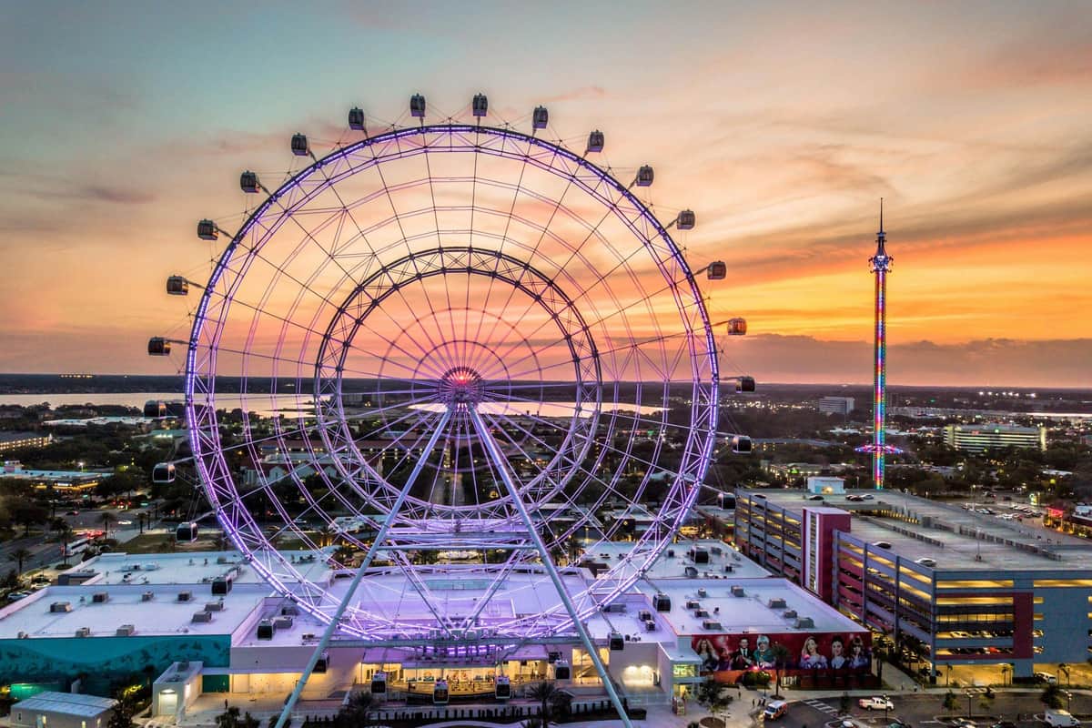 The Florida Pass A large ferris wheel lit with purple lights at sunset, with a cityscape and a tall illuminated tower in the background.