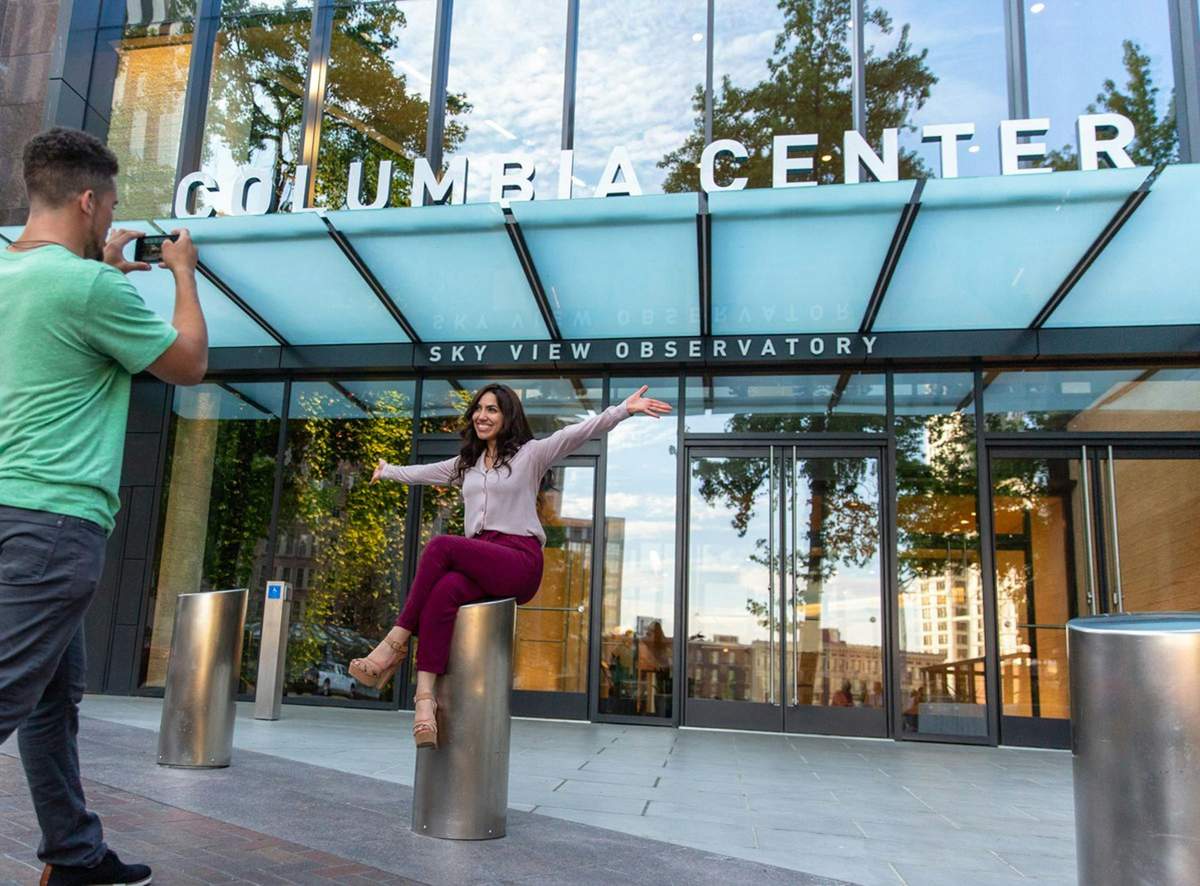 Sky View Observatory: Standard Admission A man takes a photo of a smiling woman sitting on a pole in front of the Columbia Center Sky View Observatory entrance.