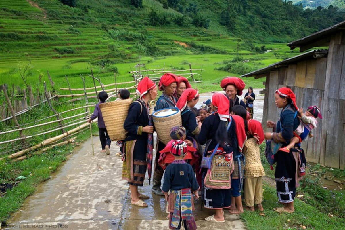 Sapa: 2-Day Guided Tour from Hanoi + Cat Cat Village & Fansipan Peak Visit Group of people, mostly women and children, wearing traditional clothing and red headscarves, gathered near wooden buildings and green fields.