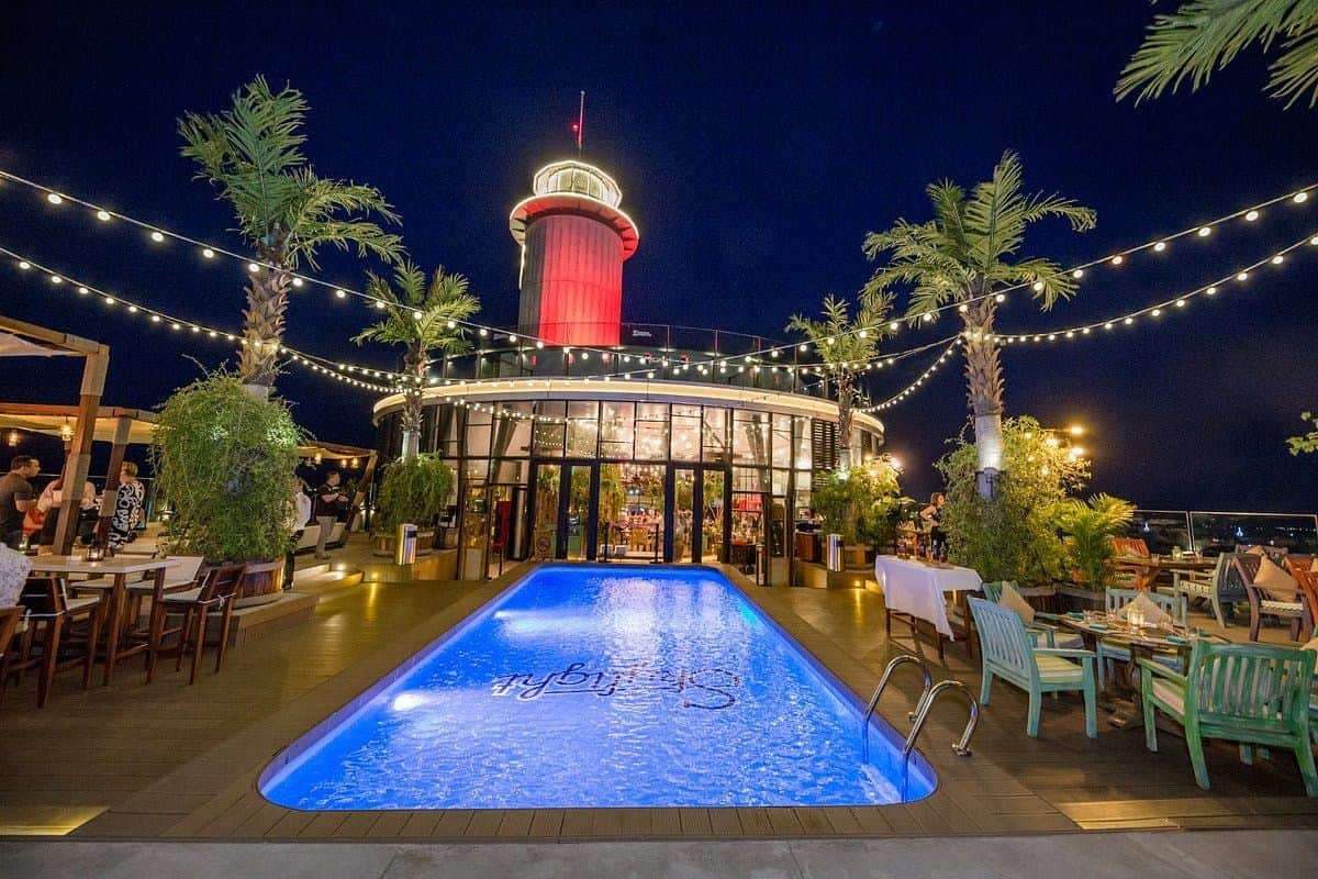 Skylight 360 Skydeck & Rooftop Beach Club Nha Trang: Entrada + Bebida Exclusiva + Petisco Uma área no terraço à noite, com uma piscina em primeiro plano, palmeiras, luzes decorativas e uma estrutura cilíndrica iluminada.