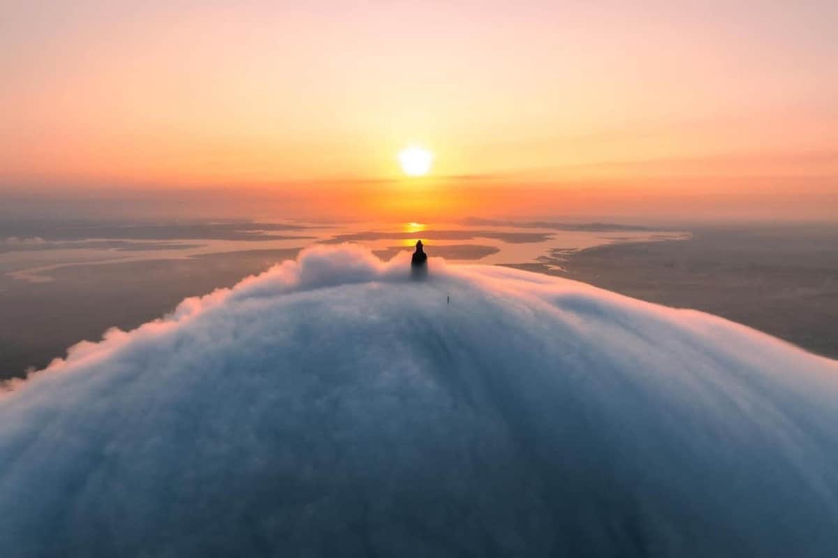 Sun World Ba Den Mountain: Roundtrip Cable Car Ride A person stands on a cloud-surrounded peak, facing a distant sunset over a calm, expansive landscape.