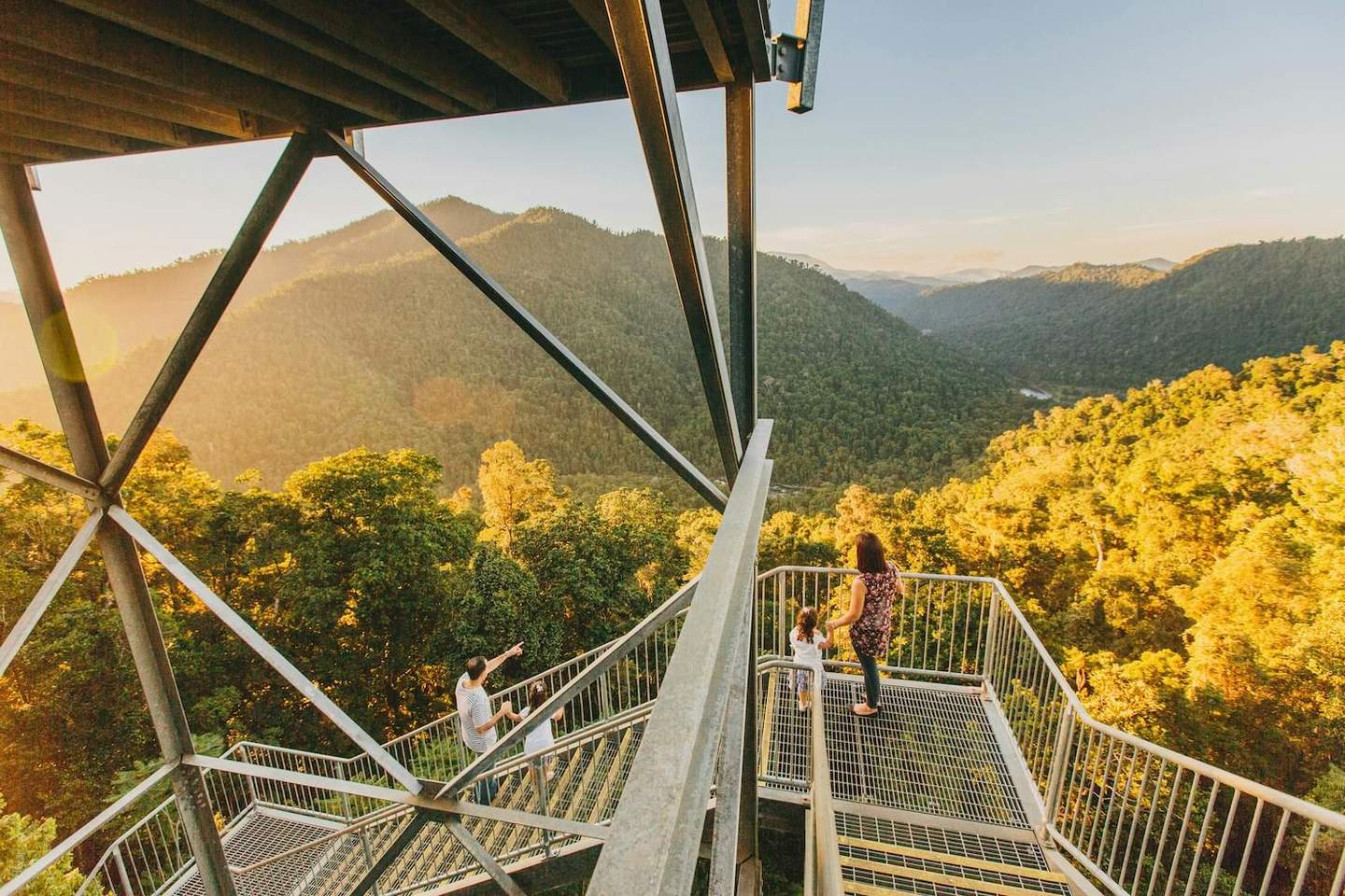 Mamu Tropical Skywalk People standing on metal platforms and stairs, admiring a sunset view of forested mountains. Metal beams frame the scene.