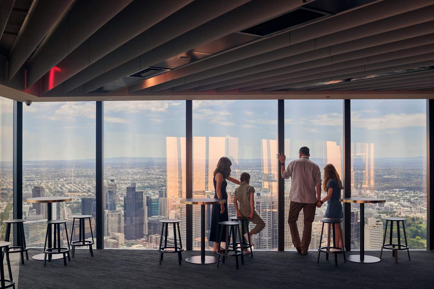 Melbourne Skydeck Four people stand by floor-to-ceiling windows in a high-rise building, overlooking a cityscape with tall buildings and scattered clouds.