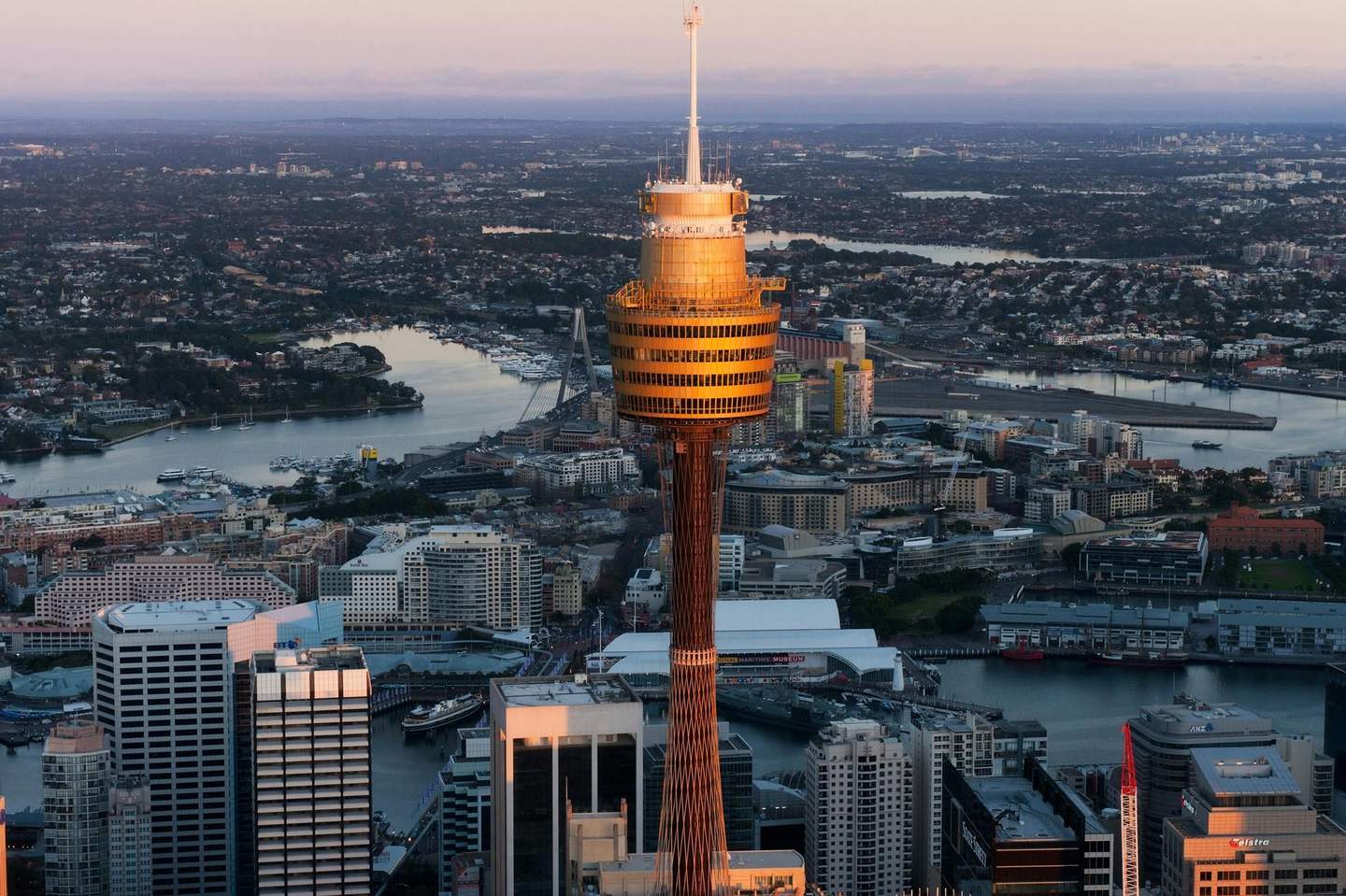 Sydney Tower Eye Aerial view of a city with a prominent yellow-gold observation tower, surrounded by buildings, waterways, and a distant bridge at sunset.