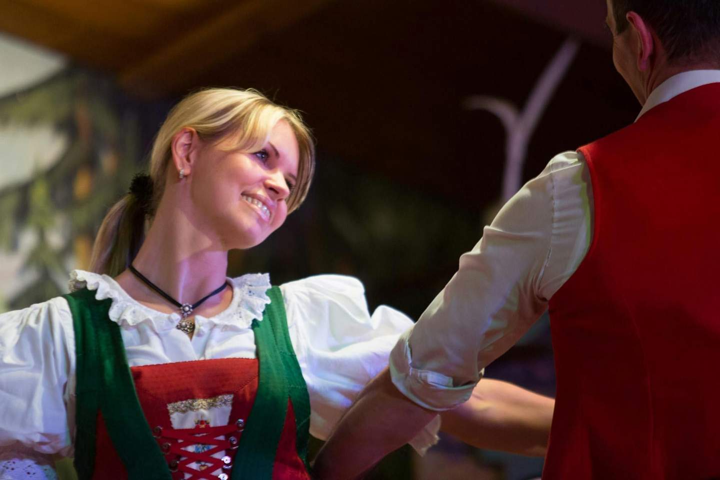 A woman in traditional attire smiles while dancing with a partner wearing a red vest.