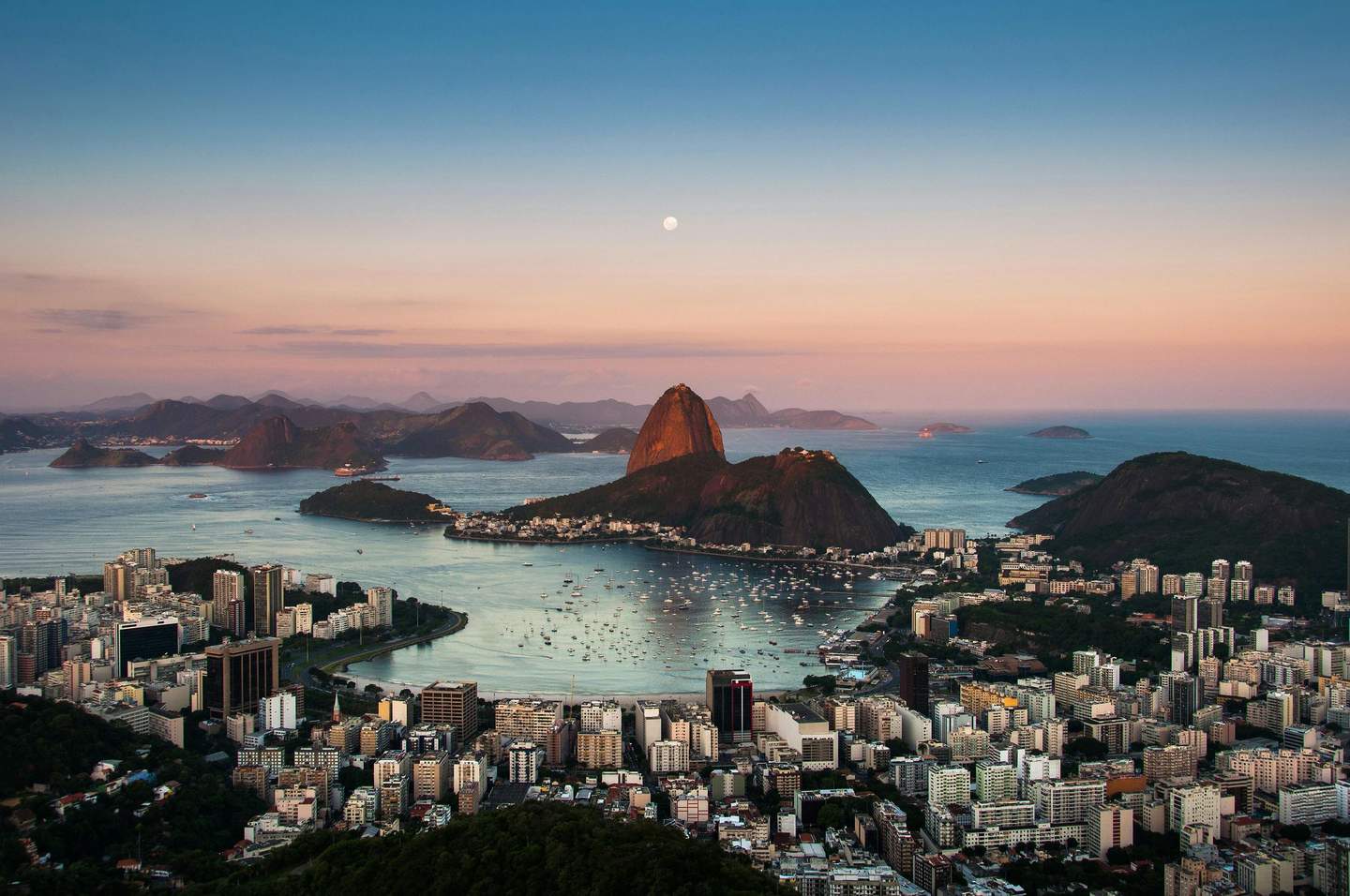 Sugarloaf Mountain A city with many high-rise buildings near a waterfront, mountains in the distance, and a moon in the sky during sunset.