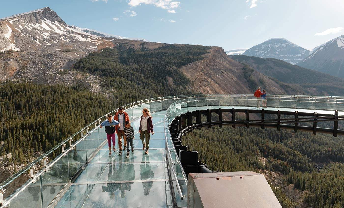 Columbia Icefield Skywalk People walking on a glass-floor observation deck in a mountainous area with forested valleys and snow-capped peaks.
