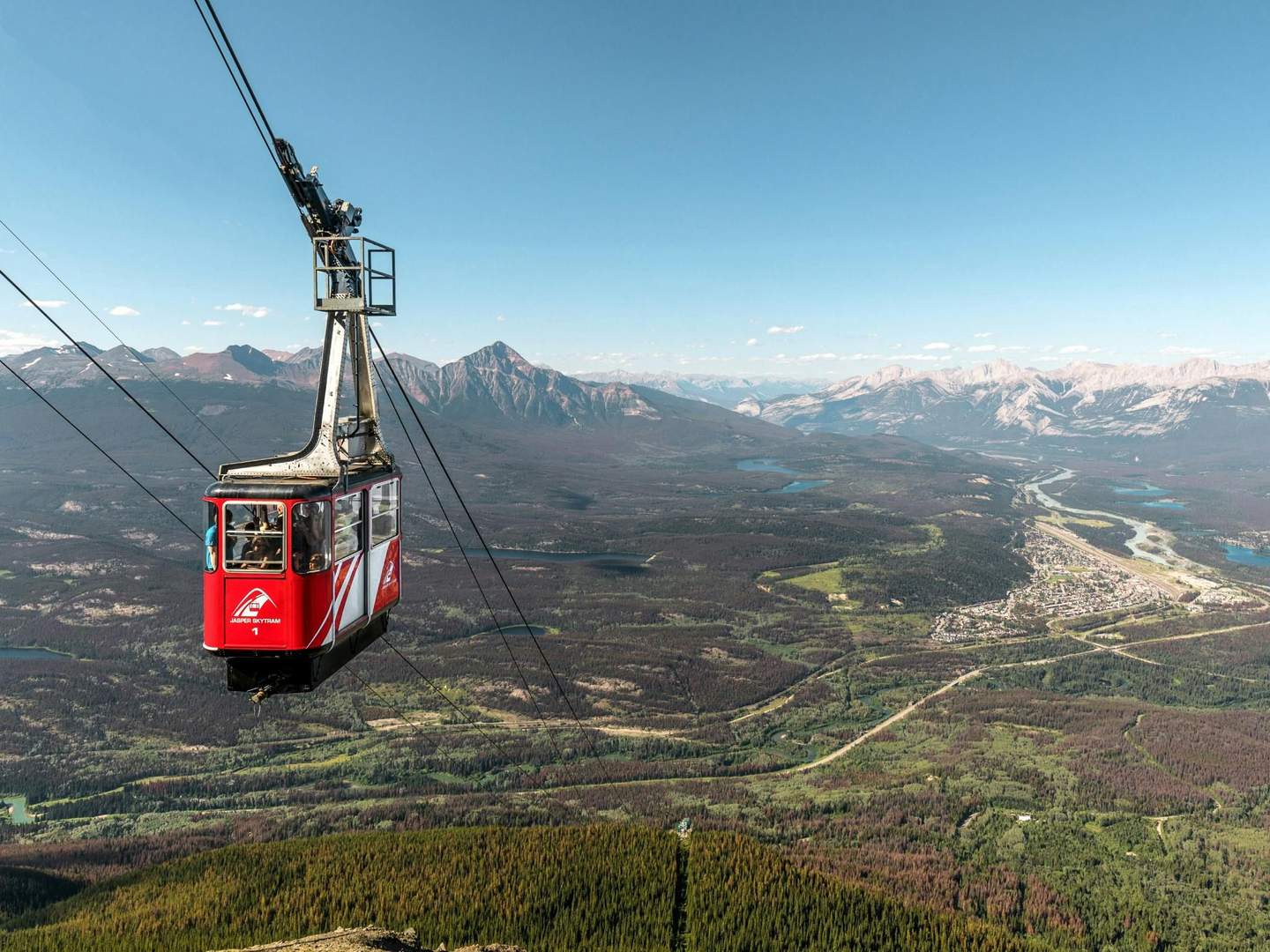 Jasper SkyTram A red cable car ascends over a vast green landscape with mountains and lakes under a clear blue sky.