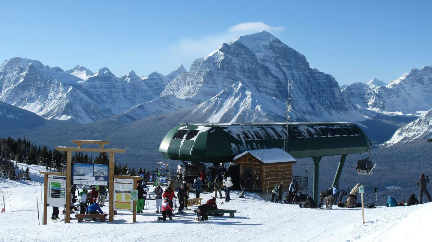 Lake Louise Ski Resort Skiers gather at a mountain ski lift station with snow-covered peaks in the background on a clear day.
