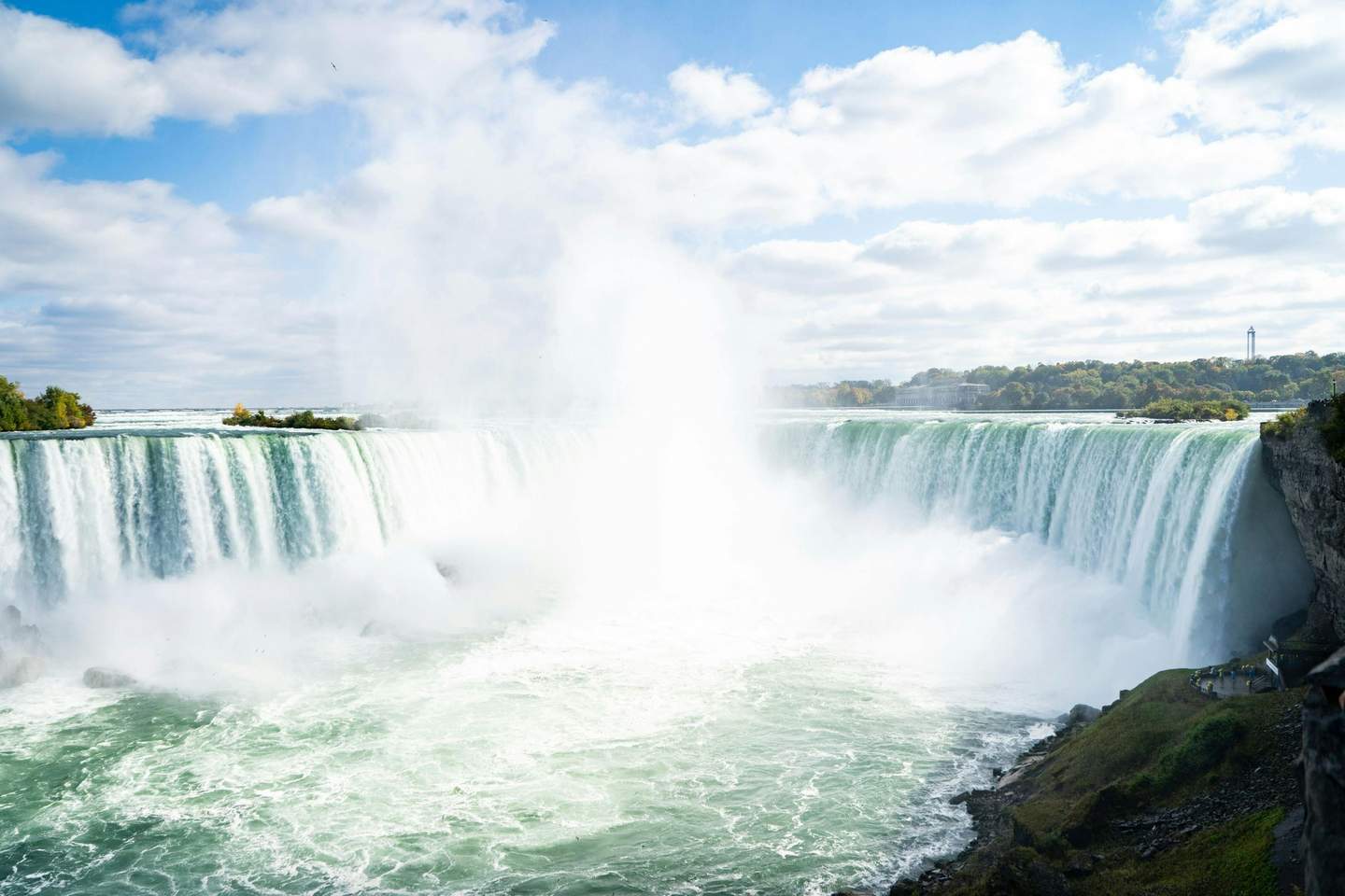 A majestic waterfall with water cascading down a cliff, creating mist, set against a partly cloudy sky and distant greenery.