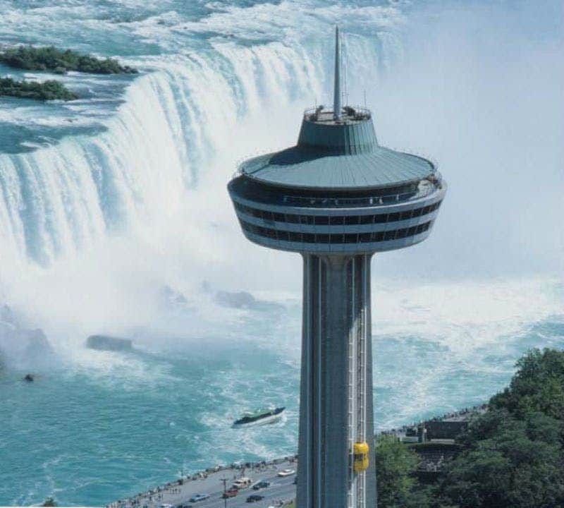 Skylon Tower A tall observation tower stands near a powerful waterfall, with a boat navigating the water below and trees surrounding the area.