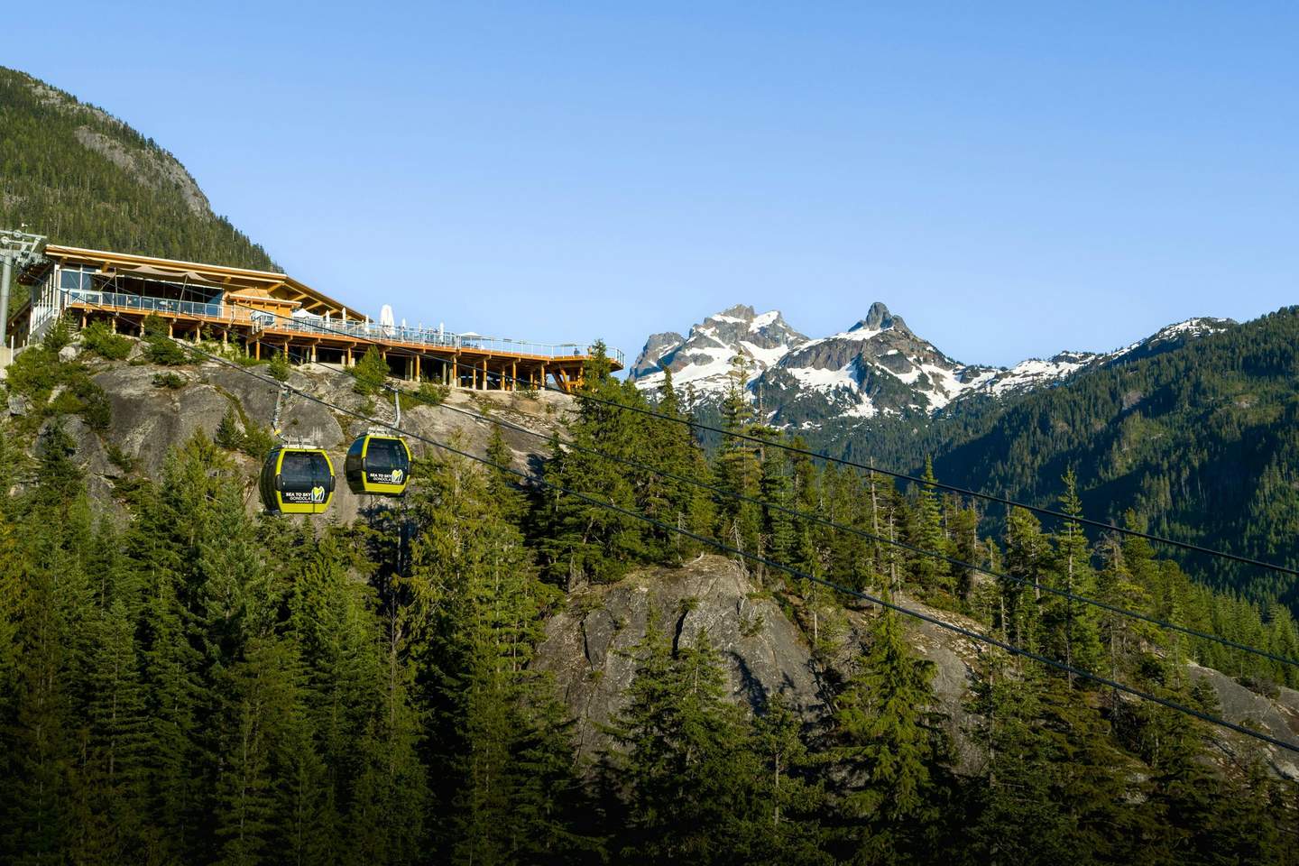 Sea to Sky Gondola Mountain gondolas in the foreground ascend toward a wooden lodge, with snow-capped peaks and forested hills in the background.
