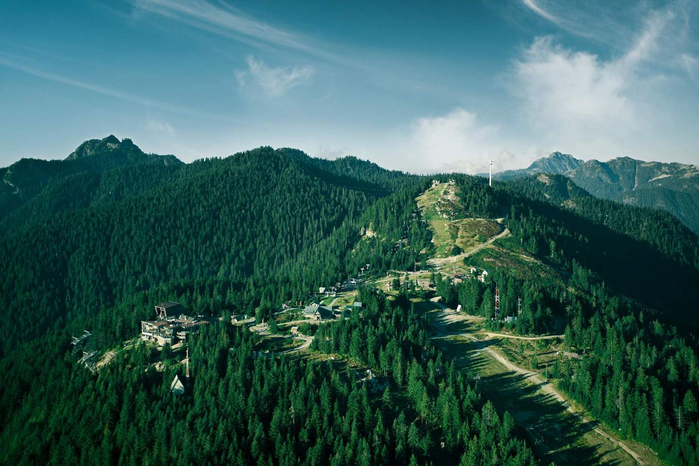 Aerial view of a forested mountain range with buildings and a winding road, under a clear sky with scattered clouds.
