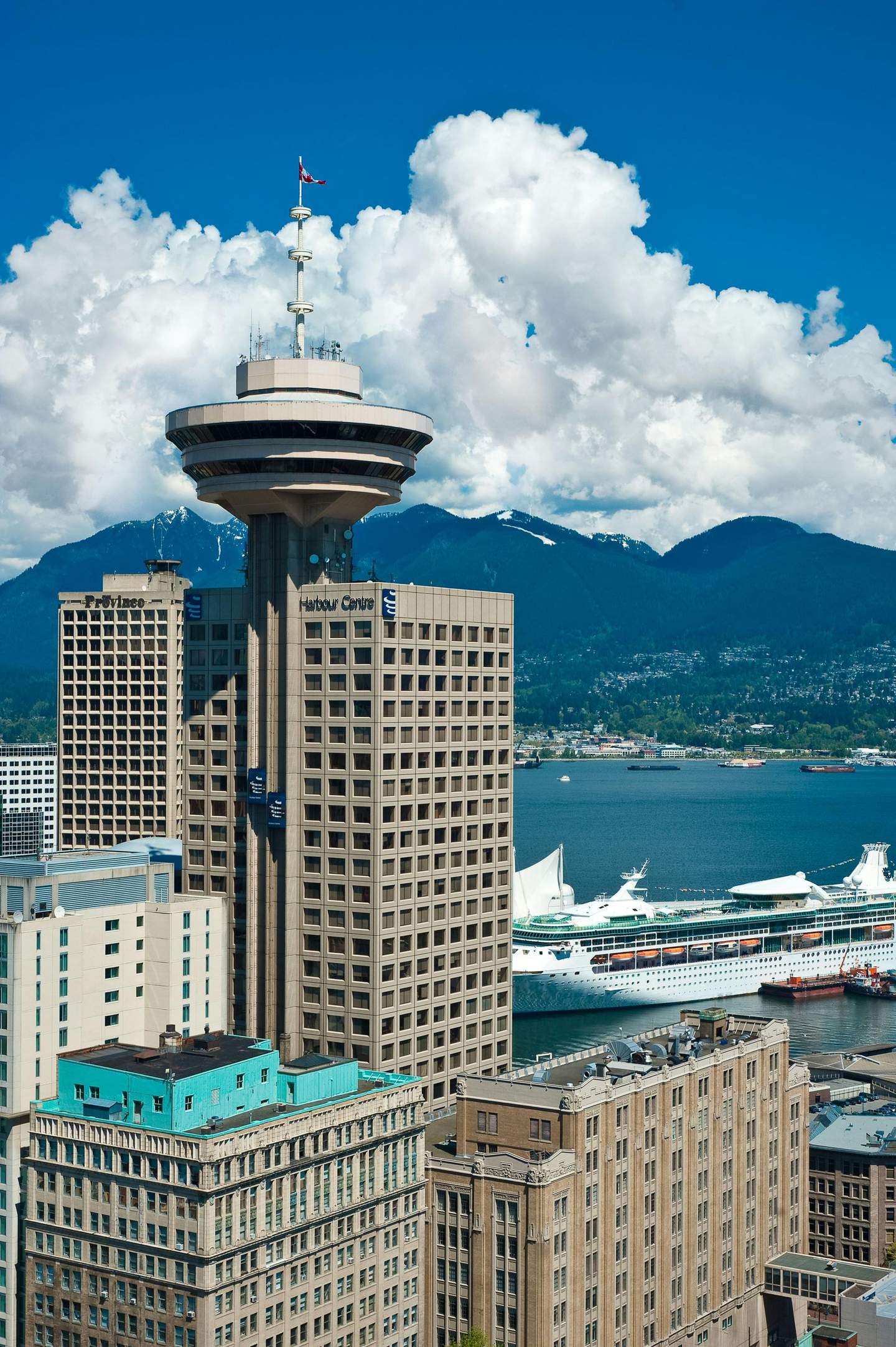 Tall buildings with Harbour Centre tower, cruise ship at the dock, ocean, mountains, and cloudy sky in the background.