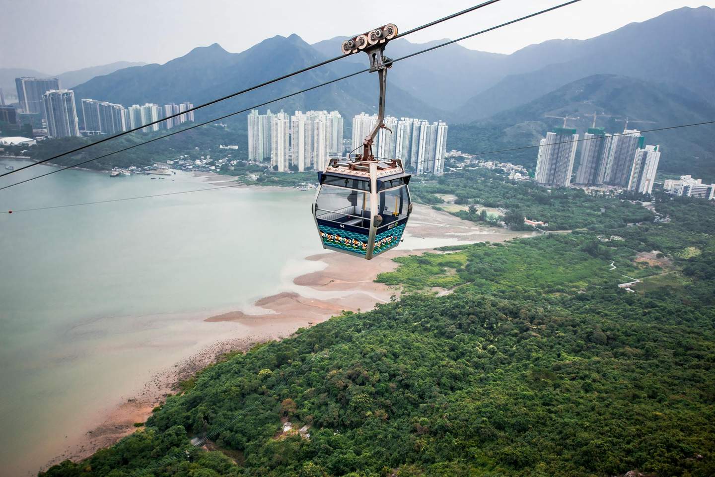 Ngong Ping Cable Car A cable car travels above a green landscape with high-rise buildings and mountains in the background near a body of water.